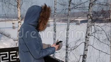 穿着蓝羽绒服的女人在冬天公园的手机里写短信。 侧视图
